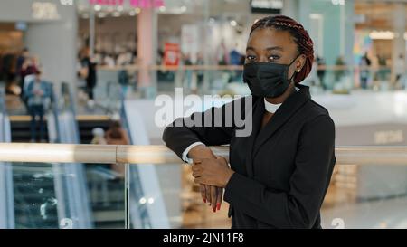 Ritratto di afro americano giovane ragazza donna acquirente consumatore indossa la maschera medica in piedi nel centro commerciale centro commerciale guardando la macchina fotografica in posa vicino ai negozi Foto Stock