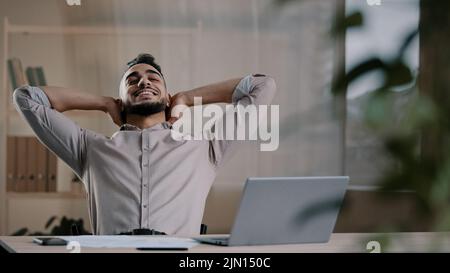 Felice soddisfatto uomo arabo lavoratore uomo d'affari ispanico ragazzo finito lavoro compito sul computer sedersi a casa ufficio mettere le mani dietro testa appoggiarsi su sedia prendere Foto Stock