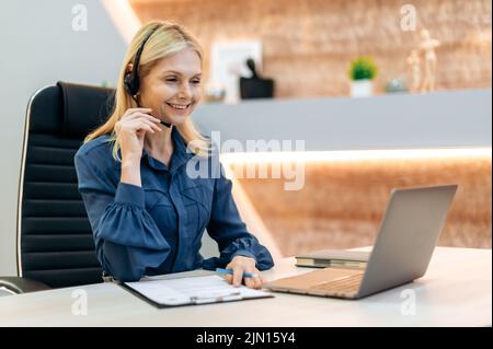 Donna d'affari caucasica di mezza età di successo amichevole, operatore di call center femminile, consulente finanziario, in abbigliamento formale, con cuffie, seduta in ufficio moderno, tiene la consultazione online con il cliente Foto Stock