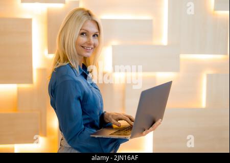 Donna caucasica matura intelligente di successo, donna d'affari, in abiti da lavoro, segretario femminile, tenendo un laptop aperto nelle mani, in piedi nel moderno ufficio, guarda la macchina fotografica, sorridente Foto Stock