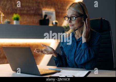 Funzionamento a ore dispari. La signora caucasica matura di affari, ceo dell'azienda, lavora straordinario, siede nell'ufficio alla sera, con un visore, conduce le trattative in linea con i colleghi o i clienti Foto Stock