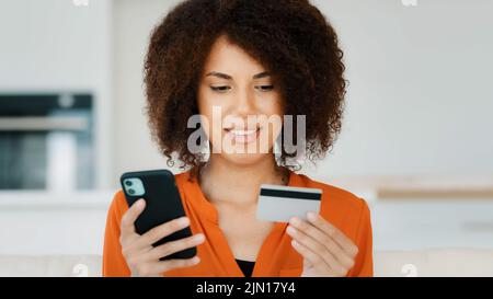 Donna africana americana che paga in linea con il servizio mobile finanziario nella cucina domestica. Ragazza biraciale con capelli ricci che tengono il telefono della carta di credito della banca Foto Stock