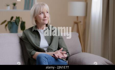 Felice sognare ispirato pensieroso anziano vecchia caucasica 60s donna guardando via nella finestra sedersi sul divano da solo pensare, anziano pensivo signora di mezza età Foto Stock
