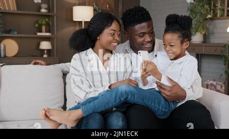 Felice famiglia africana genitori con carino schoolgirl bambino rilassarsi sul divano utilizzando divertente smartphone app lettura scherzo online ridendo divertirsi con Foto Stock