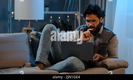 Uomo d'affari arabo indiano ispanico uomo ported utente che indossa occhiali seduti sul divano notte a tarda ora con difficoltà computer portatile in linea Foto Stock