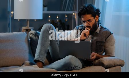 Uomo d'affari ragazzo arabo indiano uomo portato utente indossare occhiali seduti sul divano di notte in ritardo con laptop difficoltà di lettura notizie problemi online Foto Stock