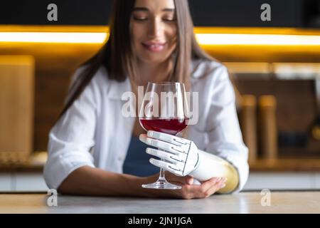 La ragazza calma e premurosa tiene un bicchiere di vino in braccio bionico a casa, donna con arto protesico artificiale. Foto Stock