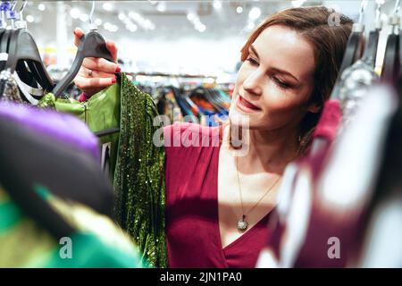 La donna graziosa sceglie i vestiti nel deposito. Ragazza acquirente guarda curiosamente al vestito. Shopping femminile Foto Stock