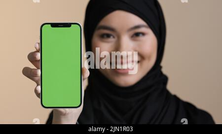 Ritratto asian ragazza musulmana donna islamica in nero hijab sorridente mostrando telefono cellulare con schermo verde tenendo smartphone guardando la fotocamera promo Foto Stock