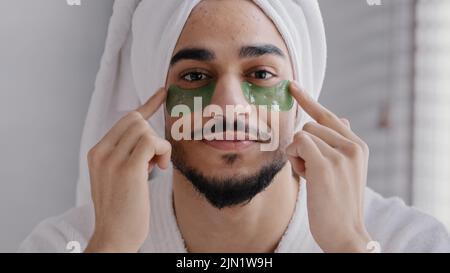 Vista frontale ritratto maschio faccia sorridente indiano uomo arabo indossare bagno asciugamano testa colla appiccicoso idrogel eye-patches procedure cosmetiche cura della pelle ragazzo Foto Stock