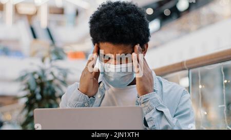 African american business man ragazzo studente indossare maschera medica lavoro con laptop in ufficio aziendale spazio tenendo testa sensazione di mal di testa sofferenza Foto Stock