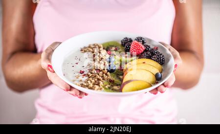 Yogurt, muesli di mora, lamponi, mirtilli, kiwi e pesche in una ciotola bianca che tiene la giovane donna. Foto Stock