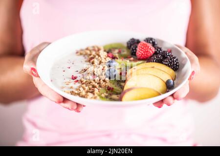 Yogurt, muesli di mora, lamponi, mirtilli, kiwi e pesche in una ciotola bianca che tiene la giovane donna. Foto Stock