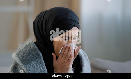 Primo piano malato sconvolto giovane ragazza in hijab starnutire naso con carta tovagliolo triste non sano musulmana che beve tè caldo che soffre di freddo stagionale Foto Stock