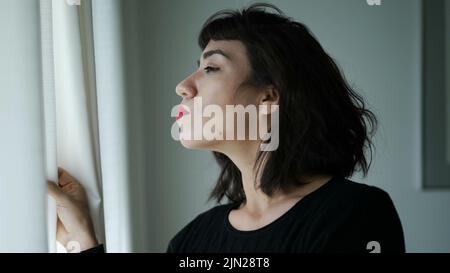 Una donna ispanica che sbirciava attraverso la finestra a tendina Foto Stock