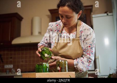 La casalinga di charme cuoca sottaceti fatti in casa, mette i vasi con cetrioli sottaceto capovolti su cassa di legno in cucina Foto Stock