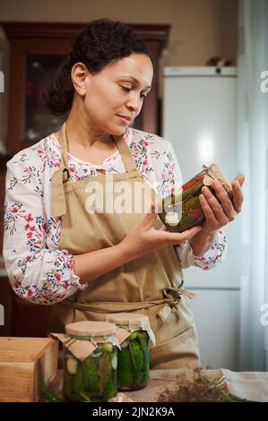 La donna graziosa guarda il vaso di peperoncino sottaceto che tiene nelle sue mani, si alza al tavolo da cucina con vasetti di sottaceti Foto Stock