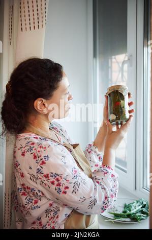 La donna matura si alza accanto alla finestra in cucina e guarda il vaso con peperoncino, in scatola secondo una ricetta di famiglia Foto Stock