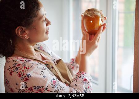 Donna Chraming si alza accanto alla finestra in cucina e guarda il vaso con confettura di pesche, in scatola secondo la ricetta di famiglia Foto Stock