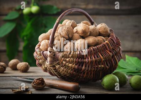 Cestino di vimini pieno di noci, noci verdi e mature, coltello su tavola di legno. Foto Stock