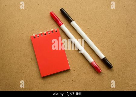 Blocco note rosso per note con fogli rossi. Spazio vuoto per le note. Mocap. Due pennarelli in rosso e nero. Su sfondo grigio ocra caldo. Caleae di festa Foto Stock