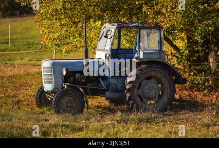 Vecchio blu, parzialmente arrugginito, trattore parcheggiato sul prato autunnale sfocato alberi sfondo Foto Stock