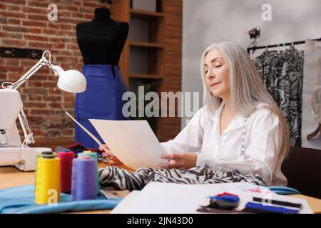 Donna con capelli grigi, stilista di moda. Guarda gli schizzi di una nuova collezione di abiti sullo sfondo di un manichino e di un cucito Foto Stock