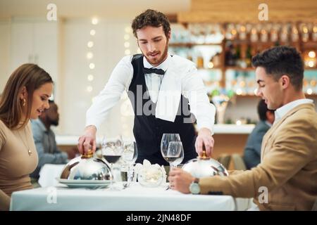 Li serve sempre con rapidità e gentilezza, un bel cameriere giovane che serve una giovane coppia in un ristorante. Foto Stock