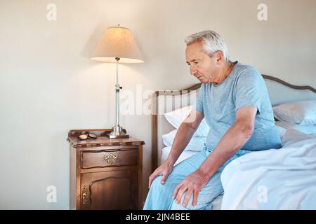 Pensare a come trascorrere la mia giornata. Un anziano seduto da solo sul bordo del letto e che guarda contemplativo a casa. Foto Stock