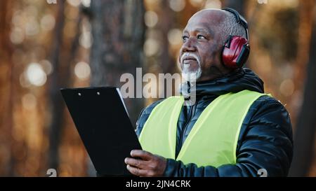 Primo piano african american uomo professionale forestale ingegnere in piedi all'aperto indossando cuffie di protezione che valutano la situazione facendo albero Foto Stock