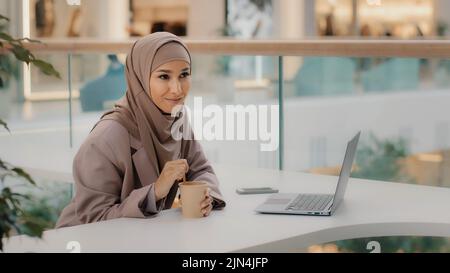 Giovane donna d'affari musulmana in hijab seduta in ufficio alla scrivania con il laptop che mescola il caffè in una tazza di carta tempo libero ragazza araba felice di leggere piacevole e-mail Foto Stock