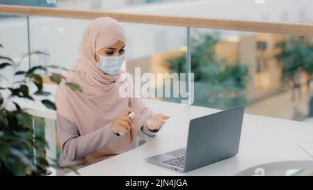 Divertente donna araba seduta in luogo pubblico usa antisettico spruzzi intorno in aria su laptop copre faccia con maschera medica ragazza musulmana paura di germi Foto Stock