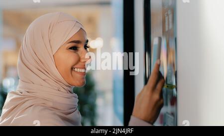 Giovane donna araba felice in hijab si trova vicino alla macchina da caffè ordini bere sorridente ragazza musulmana facendo ordine su automaton self-service paga per il servizio Foto Stock