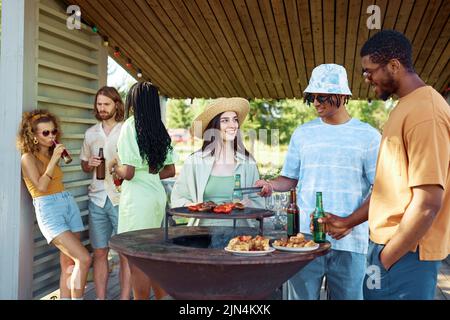 Gruppo di amici diversi che grigliano carne e verdure mentre si divergono barbecue all'aperto in estate Foto Stock
