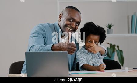 Felice afroamericano maturo padre prendersi cura sorride aiutando bambina scolaretta con studio lavoro mostrando pollici su gesto di approvazione utilizzando Foto Stock