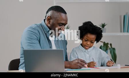 Adulto padre casa insegnante aiutare figlia piccola a fare i compiti sedersi alla scrivania in camera studio remoto utilizzando laptop uomo spiegare capretto ragazza compito africano Foto Stock