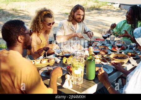 Gruppo di persone diverse che si godono la cena insieme all'aperto in estate seduti a tavola con deliziosa festa fatta in casa Foto Stock