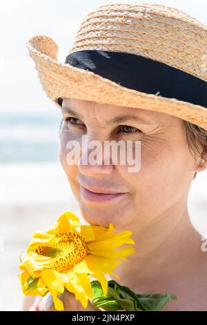 Donna in un cappello di paglia con un girasole guarda al lato, girando la faccia al vento forte, contro sfondo sfocato del mare. Foto Stock