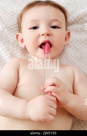 Il bambino felice del toddler impara a spazzolarsi i suoi denti mentre si trova sul suo letto di casa. Il bambino pulisce la bocca con uno spazzolino nelle mani. Bambino di un anno Foto Stock