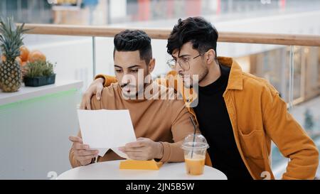 Due amici studenti seduti in un caffè giovane uomo preoccupato che riceve una lettera di lettura cattive notizie sconvolto sente triste fallimento esame licenziamento perdita di problemi finanziari Foto Stock