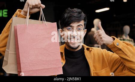 Giovane bel ragazzo gioioso si erge in boutique tenendo borse di carta punti per il buon acquisto felice uomo elegante in negozio di abbigliamento mostrando pollice su gesto Foto Stock