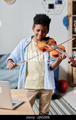 Ragazzo africano che guarda il monitor sul portatile sul tavolo e suona lo strumento musicale durante la lezione online a casa Foto Stock