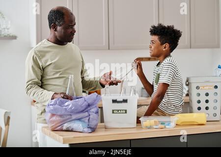 Papà africano che dà bottiglie di vetro a suo figlio per ordinarle in contenitori mentre si trovano in cucina Foto Stock