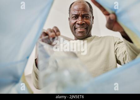 Vista ad angolo basso dell'uomo africano sorridente che lancia una bottiglia di plastica nel cestino Foto Stock