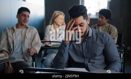 Triste frustrato studente arabo seduto in classe alla scrivania solo che soffre di maltrattamenti da parte dei compagni di classe discriminazione razziale si sente vittima di bullismo Foto Stock