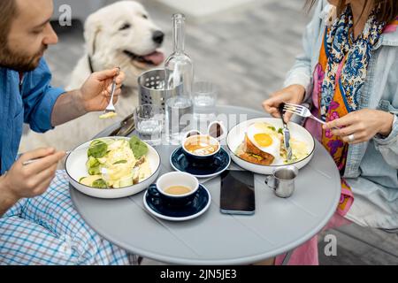 L'uomo e la donna alla moda hanno una colazione mentre si siede con il cane al caffè all'aperto Foto Stock