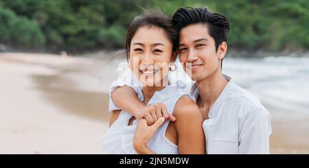 Felice sorridente Asian 30s coppia in rapporto guardando la macchina fotografica sulla spiaggia in estate. Una coppia romantica che indossa una camicia bianca e un vestito. Foto Stock