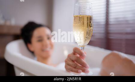 Fuori fuoco giovane donna felice e rilassata sdraiata in bagno di schiuma in vetro champagne divertente ragazza giocosa soffiando sulla schiuma in bagno godendo flavorous Foto Stock