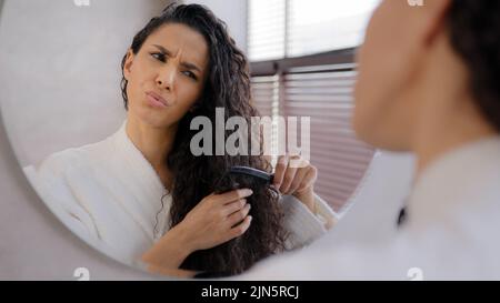 Primo piano giovane donna che guarda nello specchio pettinare i capelli in bagno infastidito triste ragazza insoddisfatto danneggiato secco aggrovigliato condizione dei capelli frustrati circa Foto Stock