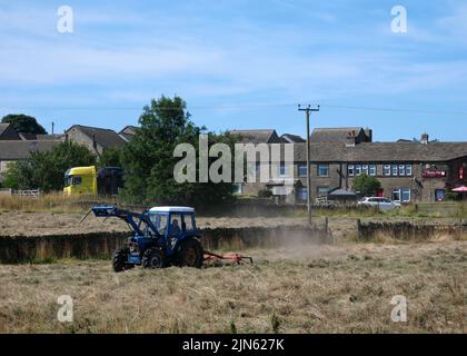Trattore blu in campo di fieno che gira l'erba in case estive sullo sfondo Foto Stock